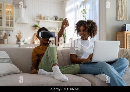 Donna afroamericana sorridente che guarda il figlio indossando un simulatore di realtà virtuale a casa Foto Stock