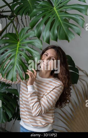 Giovane donna felice positiva che tocca la foglia di Monstera, trascorrendo del tempo con le piante nel giardino di casa Foto Stock