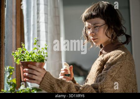 Donna millenaria che appannano le piante domestiche a casa, utilizzando spray per piante per insetti Foto Stock