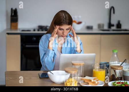 Donna lavoratrice stanca e esausta con mal di testa che lavora su un computer portatile seduto al tavolo in cucina. Foto Stock