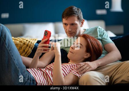 Coppia di mezza età soddisfatta e ben riposata che si rilassa sul divano a casa riposando con lo smartphone. Foto Stock