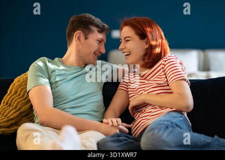 Donna felicissima che ride sinceramente con un ragazzo sorridente che si tiene per mano, sedersi sul divano a casa Foto Stock