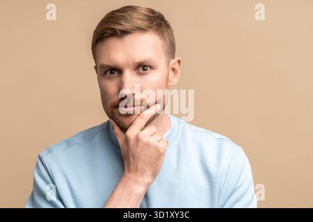 Un uomo barbato di mezza età attento e serio che tocca il mento e guarda la fotocamera su uno sfondo beige Foto Stock