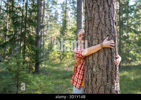 Uomo rilassato che abbraccia il tronco di albero coccolato sulla natura nella foresta con gli occhi chiusi che proteggono l'ambiente. Foto Stock
