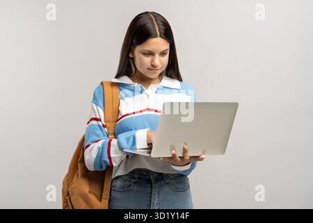 Studentessa interessata adolescente con zaino che guarda lo schermo del notebook su sfondo grigio. Foto Stock