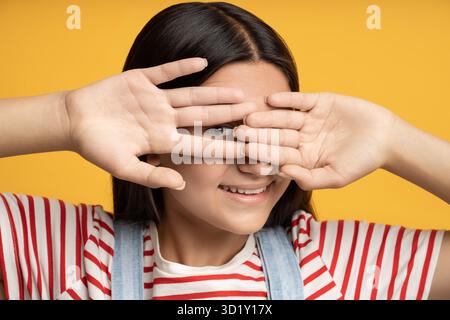 Ritrarre una ragazzina sorridente che si nasconde tra le mani con un occhio su sfondo giallo. Foto Stock