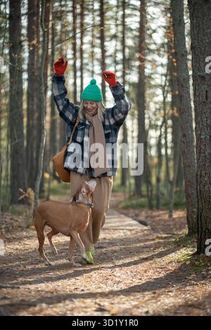 Donna allegra che gioca con il cane da salto e corsa mentre cammina nella pineta in una soleggiata giornata autunnale Foto Stock