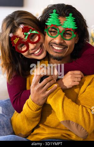 Coppia asiatica che si abbraccia sul divano in soggiorno indossando occhiali da vista festivi e tenendo in mano lo smartphone Foto Stock
