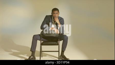Posa di un uomo adulto che indossa una tuta navy appoggiato in avanti su una sedia pieghevole in studio, con una posa attenta Foto Stock