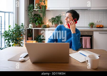 Una donna matura sorridente ama le chiamate tramite smartphone mentre mette in pausa gli studi online sul laptop. elearning remoto Foto Stock
