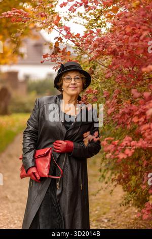 Una donna sorride mentre passeggia in un vivace parco autunnale. Indossa un cappotto di pelle nera e guanti rossi, tenendo in mano una borsa rossa. Foglie colorate surroun Foto Stock