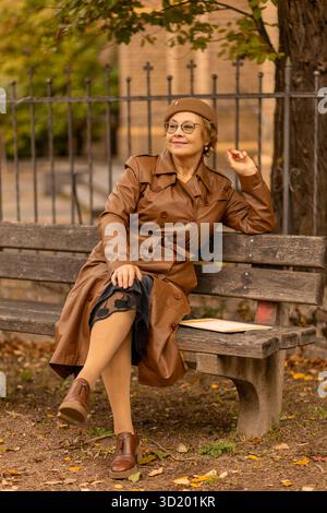 Una donna siede comodamente su una panchina in un parco, indossando un cappotto marrone e occhiali. Sorride mentre si rilassa tra il fogliame autunnale, godendosi la tranquillità Foto Stock