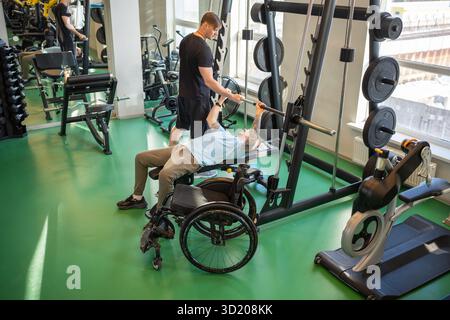 L'uomo disabile in sedia a rotelle solleva il barbell, si concentra sul movimento delle braccia mentre l'allenatore fornisce supporto in palestra. Recupero muscolare post lesione rafforza Foto Stock