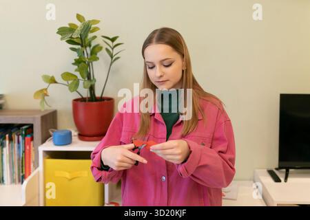 Giovane donna con una giacca rosa che taglia carta in una stanza luminosa con piante e decorazioni colorate Foto Stock