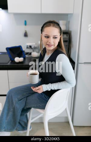 Giovane donna che si gode un caffè mentre siede in una cucina moderna durante una mattinata di sole Foto Stock