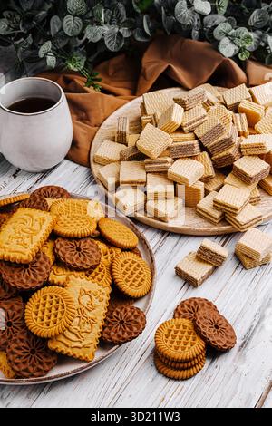 Biscotti e cialde assortiti esposti su un tavolo di legno con tazza da caffè e foglie verdi Foto Stock