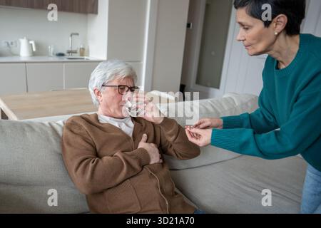 La moglie caregiving offre pillole e acqua per il cuore e la pressione sanguigna a marito debole, aiutare l'uomo malato Foto Stock