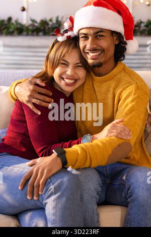 Coppia variegata che indossa il cappello di Babbo Natale e la fascia di corna che si abbraccia sul divano beige accanto al camino Foto Stock