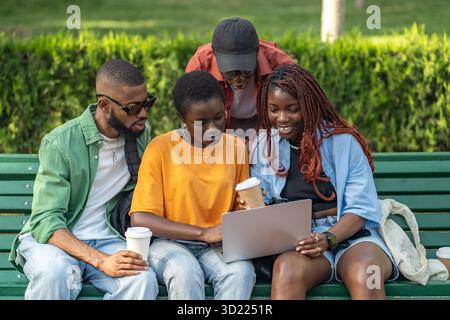 Allegro gruppo di amici afroamericani che bevono caffè e lavorano su computer portatili nel parco verde Foto Stock