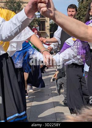 Persone in costumi tradizionali che si tengono per mano, eseguono una danza popolare su una strada lastricata durante un vivace festival all'aperto Foto Stock