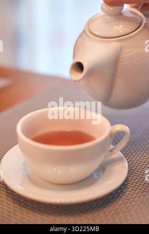 Teiera bianca che versa il tè appena preparato in una tazza bianca sul tavolo con un morbido composto Foto Stock