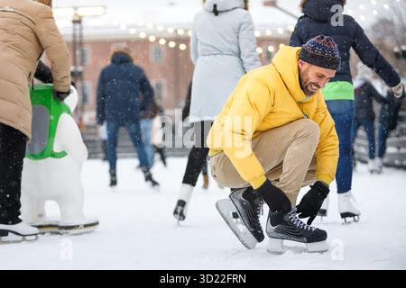 Uomo di mezza età caucasico che lega pattini neri sulla pista di pattinaggio su ghiaccio in giornate invernali innevate all'aperto Foto Stock