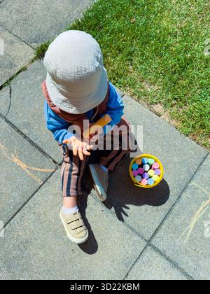 Un bambino si siede gioiosamente su un pavimento caldo, giocando con gessetti colorati ed esprimendo creatività in un ambiente all'aperto soleggiato, circondato da erba verde a. Foto Stock