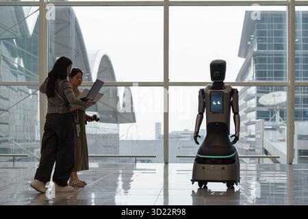 Hong Kong, Cina - 20 settembre 2025: Due donne stanno in piedi e tengono in mano lapplani vicino a un robot di servizio nell'aeroporto internazionale di Hong Kong. Foto Stock