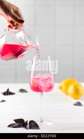 La mano di una donna versa una rinfrescante limonata di basilico rosso da una caraffa in un bicchiere. Bevande estive. Foto Stock
