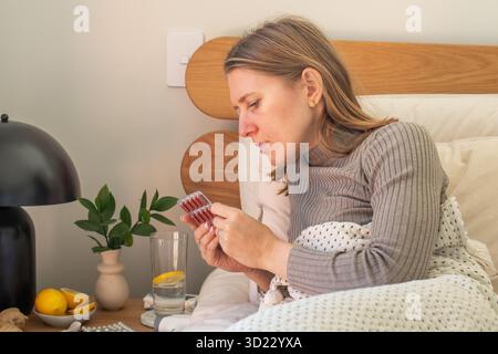 Una donna malata studia medicine mentre è sdraiata a letto. Farmaci per l'influenza e le infezioni respiratorie. Foto Stock