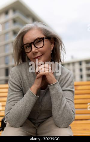 Donna anziana seduta comodamente su una panchina in un parco cittadino mentre sorride dolcemente alla telecamera durante il pomeriggio Foto Stock