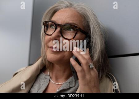 Una donna anziana sorride mentre parla sul suo smartphone in un ambiente urbano moderno durante il pomeriggio Foto Stock