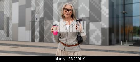 Donna che tiene in mano caffè e smartphone mentre si trova in un ambiente urbano moderno durante il giorno Foto Stock