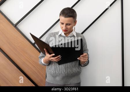 La donna che si trova in ufficio con una cartella per i documenti guarda il rapporto contro il muro Foto Stock