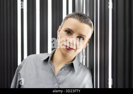 Ritratto di un'elegante donna dai capelli corti con una camicia grigia contro una parete nera con lampade retroilluminate Foto Stock