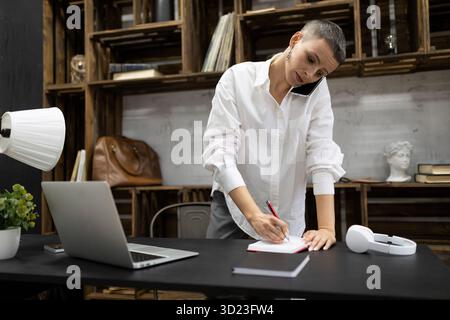 Una donna che lavora in ufficio parla al telefono e prende appunti in un notebook mentre si trova vicino al luogo di lavoro in ufficio Foto Stock