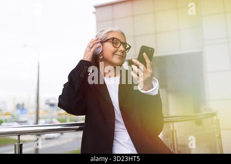 Elegante donna anziana ascolta la musica con cuffie di grandi dimensioni con un telefono cellulare e balla sullo sfondo di un moderno edificio Foto Stock