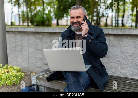 Imprenditore uomo maturo che lavora online su un notebook in un parco in una città che parla al telefono con un sorriso Foto Stock