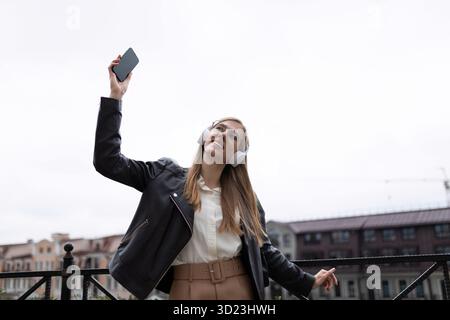 Una giovane donna felice con un telefono cellulare in mano e le cuffie balla sullo sfondo del panorama della città Foto Stock