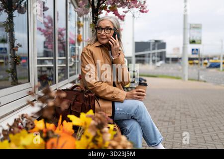Donna anziana che parla al telefono seduto su una panchina vicino al negozio Foto Stock