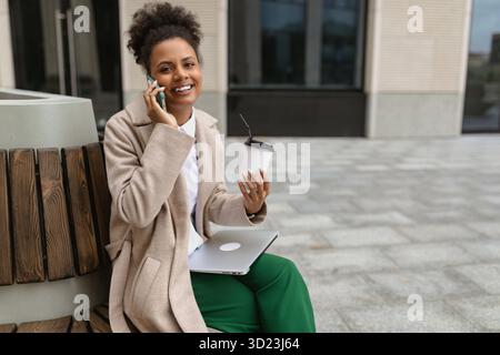 Una giovane donna afroamericana seduta vicino al business center durante la pausa pranzo con un computer portatile e un bicchiere di caffè parla con lei Foto Stock