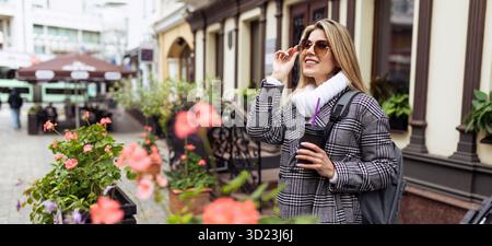 Una donna caucasica matura dall'aspetto europeo raddrizza gli occhiali da sole con una tazza di caffè in mano durante una passeggiata turistica Foto Stock