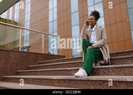 Elegante donna afroamericana che parla al telefono cellulare mentre si siede sui gradini dell'edificio degli uffici Foto Stock