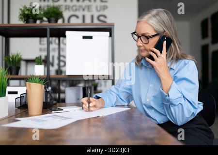 Donna d'affari adulta che lavora in ufficio e parla al telefono cellulare. Concetto di gestione fiscale fattura aziendale Foto Stock