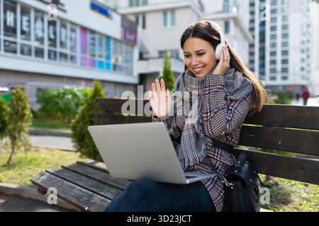 Giovane donna con un notebook in ginocchio in cuffia all'esterno sorridente con piacere e con le mani in alto Foto Stock