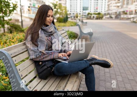 Giovane freelance di successo che lavora su un notebook sullo sfondo di un parco cittadino con caldi abiti autunnali Foto Stock