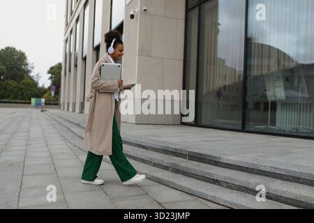 Una studentessa afroamericana con le cuffie e un portatile nelle sue mani va nell'edificio degli uffici, concetto di successo Foto Stock