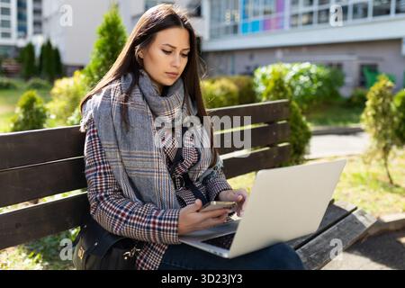 Una giovane donna con abiti autunnali lavora intensamente su un notebook mentre è seduta su una panchina all'esterno Foto Stock