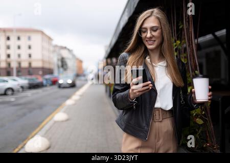 Una giovane donna elegante con un telefono tra le mani e un bicchiere di caffè sta camminando per strada Foto Stock