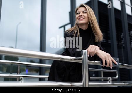 Business di successo donna bionda di mezza età con un sorriso guarda in lontananza sullo sfondo di un edificio di uffici Foto Stock